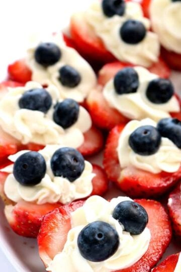 Red, white and blue strawberry cheesecake bites made with fresh strawberries, creamy cheesecake filling, and blueberries on a white serving platter.