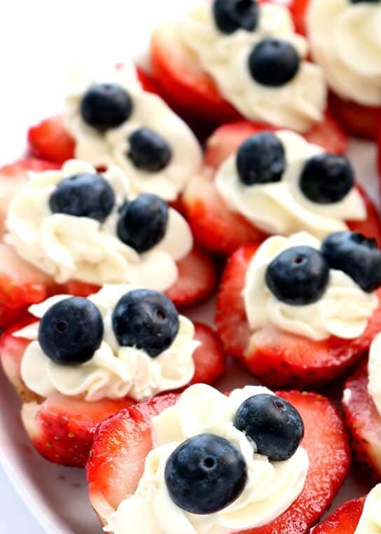 Red, white and blue strawberry cheesecake bites made with fresh strawberries, creamy cheesecake filling, and blueberries on a white serving platter.