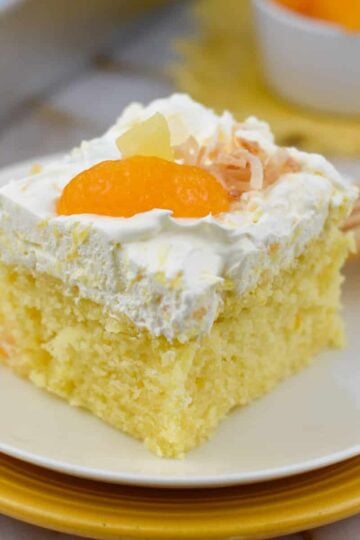 A slice of Pig Pickin' Cake topped with fluffy pineapple whipped frosting and mandarin oranges on a white dessert plate. This classic Southern dessert starts with a moist mandarin orange cake made from a box cake mix and is finished with a light, fruity topping, making it the perfect summer dessert recipe for potlucks, cookouts, and family gatherings.