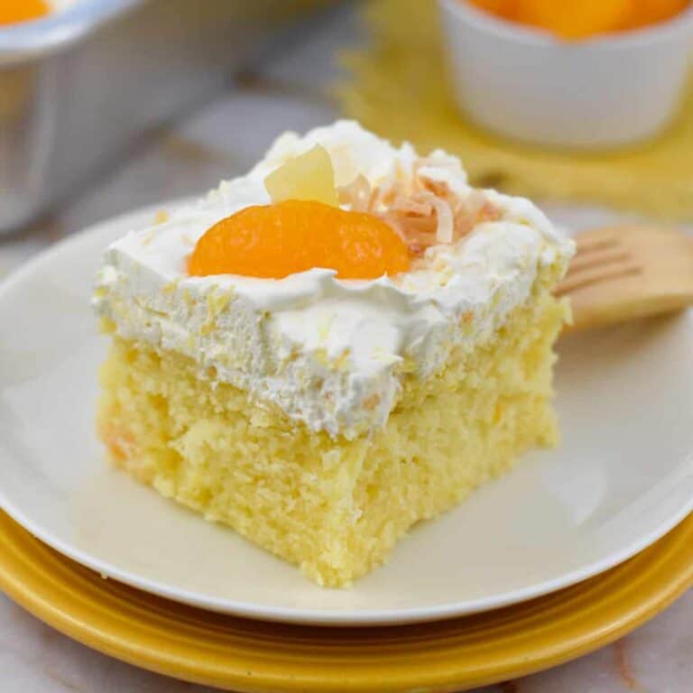 A slice of Pig Pickin' Cake topped with fluffy pineapple whipped frosting and mandarin oranges on a white dessert plate. This classic Southern dessert starts with a moist mandarin orange cake made from a box cake mix and is finished with a light, fruity topping, making it the perfect summer dessert recipe for potlucks, cookouts, and family gatherings.
